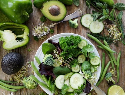 sliced broccoli and cucumber on plate with gray stainless steel fork near green bell pepper, snowpea, and avocado fruit